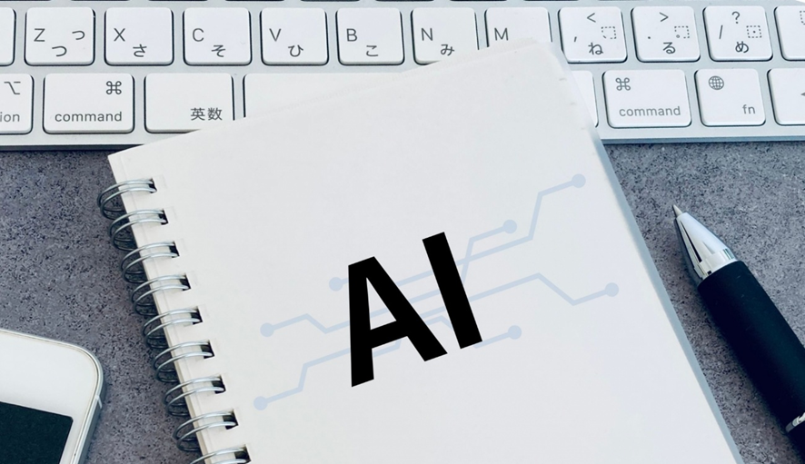 AIと書かれたノートがキーボードと筆記具に囲まれたデスク環境の写真